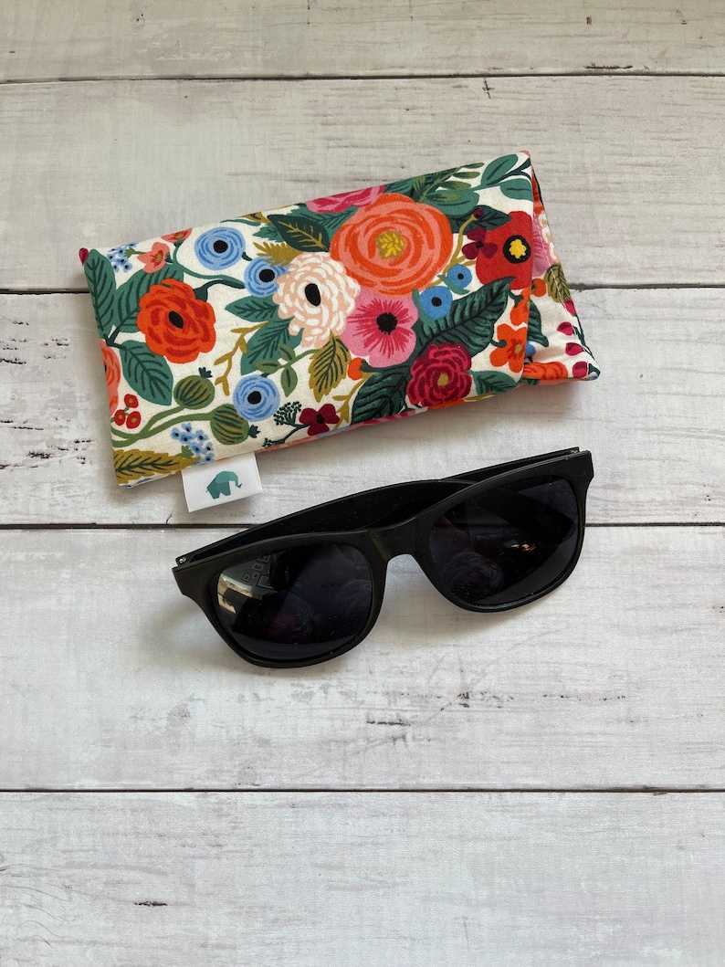 May include: A floral patterned eyeglass case with a white background and colourful flowers. The case is lying on a white wooden surface next to a pair of black sunglasses.
