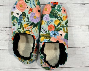 May include: A pair of floral-patterned slippers. The slippers feature a light blue background with a colorful floral design in shades of pink, purple, yellow, and green. The interior is white, and the edges are trimmed with black fabric.