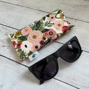 Puede incluir: Gafas de sol negras con lentes oscuras sobre una superficie de madera blanca junto a un estuche para gafas con estampado floral. El estuche presenta un patrón de flores rosas, rojas y verdes sobre un fondo crema. Una pequeña etiqueta con un logotipo de elefante está adjunta.