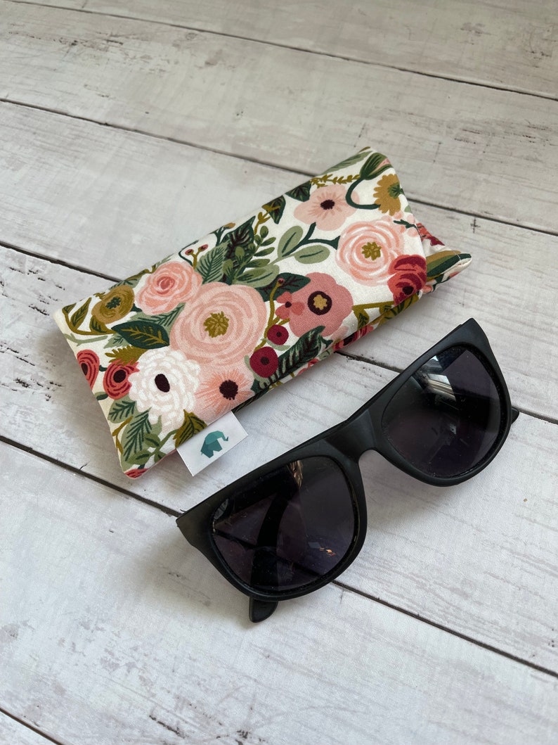May include: Black sunglasses with dark lenses rest on a white wooden surface next to a floral print glasses case. The case features a pattern of pink, red, and green flowers on a cream background. A small tag with an elephant logo is attached to the case.
