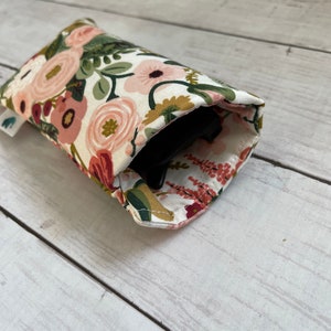 May include: A floral print glasses case featuring a pattern of pink, peach, and burgundy flowers with green leaves on a white background. The case is open, revealing a black object inside. The case is made of fabric.