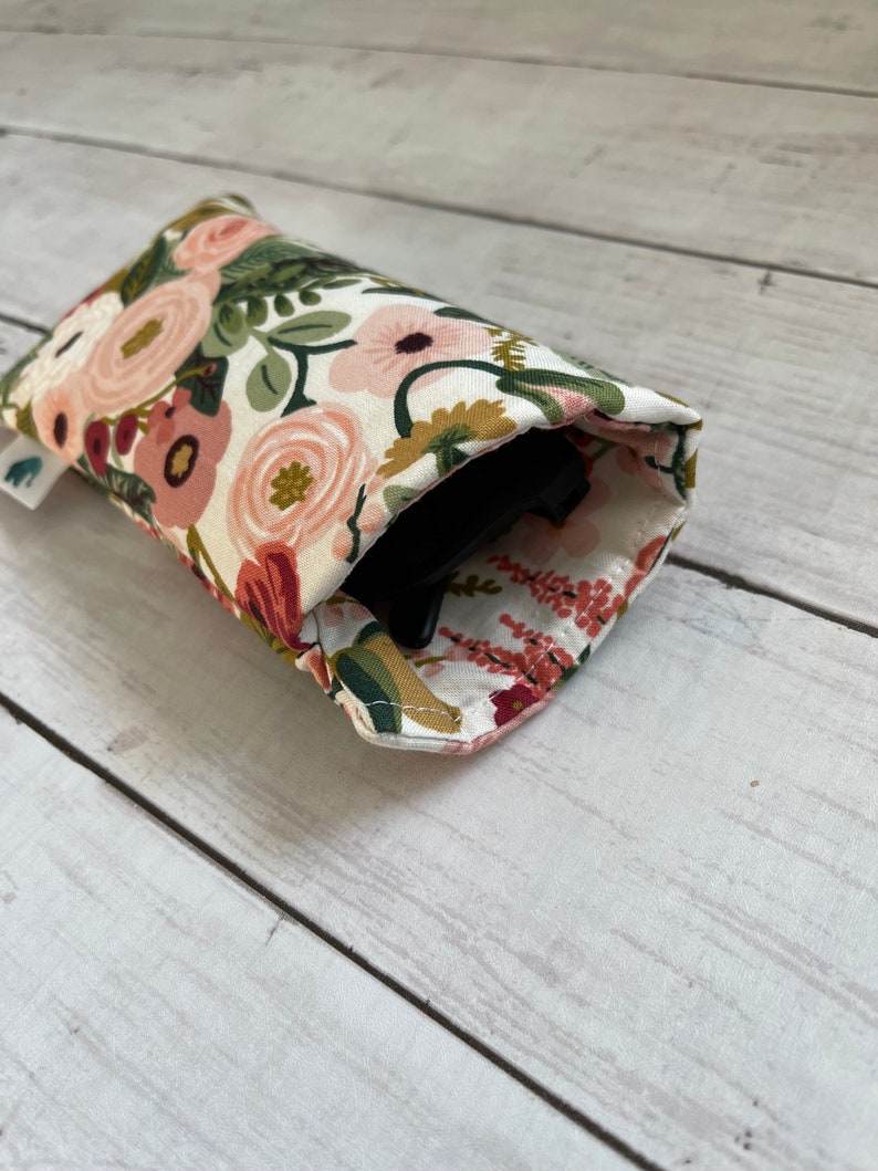May include: A floral print glasses case featuring a pattern of pink, peach, and burgundy flowers with green leaves on a white background. The case is open, revealing a black object inside. The case is made of fabric.