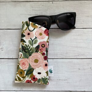 May include: Black sunglasses rest in a floral print glasses case. The case features a white background with pink, red, and green floral patterns. A small white tag with a green logo is attached to the case.