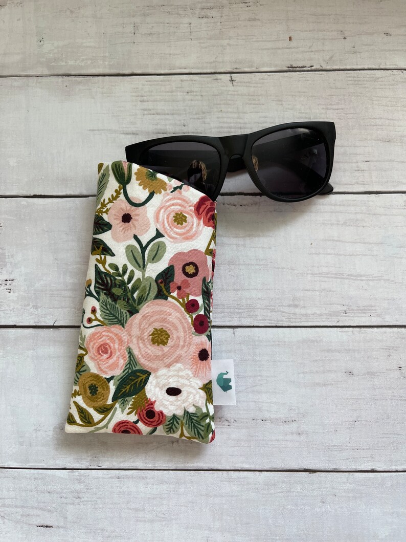 May include: Black sunglasses rest in a floral print glasses case. The case features a white background with pink, red, and green floral patterns. A small white tag with a green logo is attached to the case.