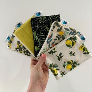 Peut inclure: Une main tient un éventail de petites pochettes zippées. Les pochettes présentent divers motifs, notamment des citrons, des motifs floraux et un fond noir avec des feuilles vertes. Chaque pochette a une petite étiquette avec un motif bleu.