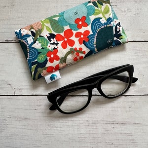 May include: A floral patterned eyeglass case with a black and white elephant logo. The case is lying on a white wooden surface next to a pair of black framed eyeglasses.
