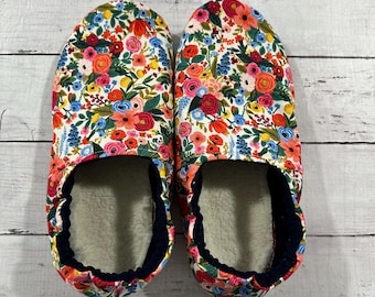 May include: A pair of floral print slippers. The slippers feature a white background with a colorful pattern of red, pink, orange, blue, and yellow flowers. The interior is lined with a soft, cream-colored material, and the edges are trimmed with navy blue fabric.