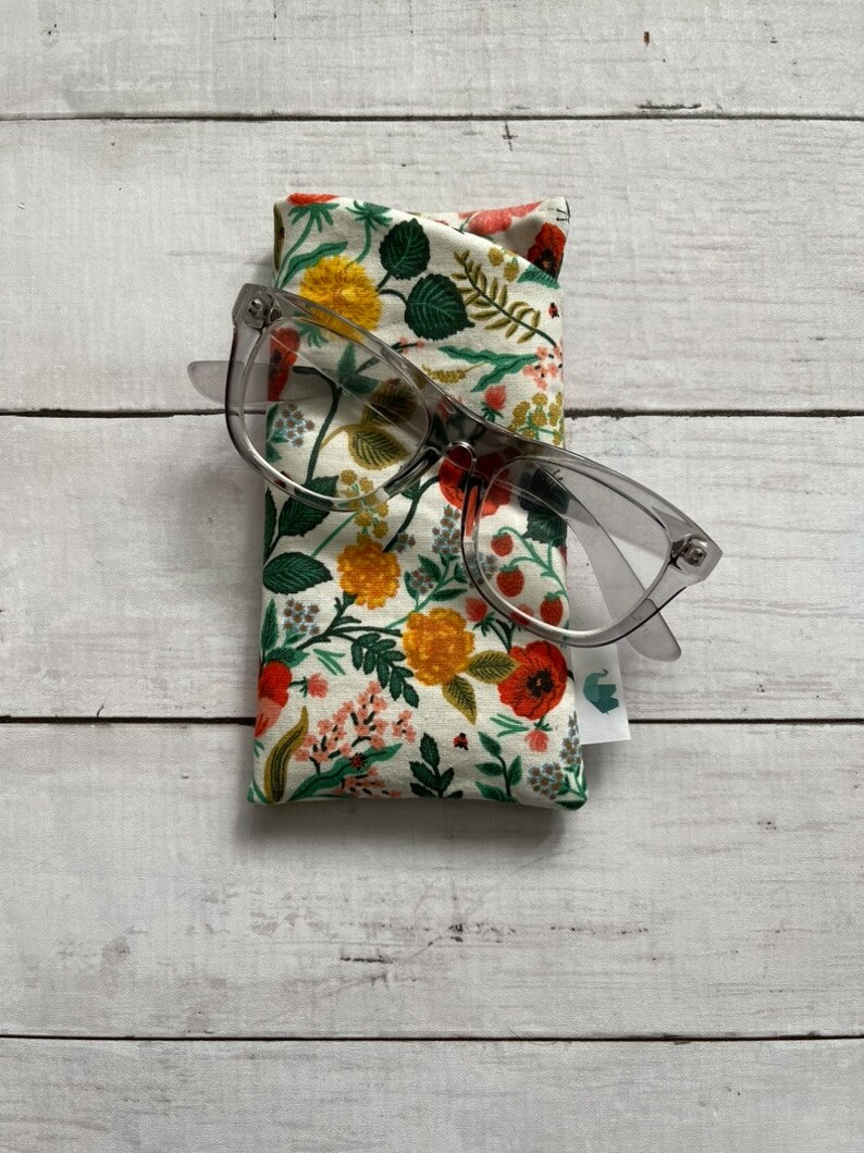 May include: A floral print glasses case with a pair of clear glasses on top of it. The case is made of fabric and has a white background with red, yellow, and green flowers.