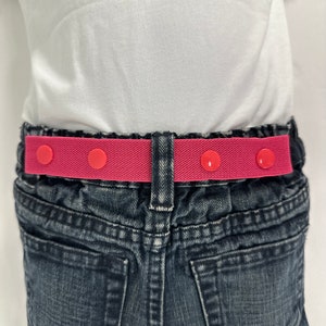 May include: A pink elastic belt with three red buttons. The belt is worn around the waist of a person wearing jeans.