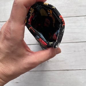 May include: A small, open fabric pouch held in a hand. The pouch is black with a colorful pattern of fruits and flowers. The interior is black. The pouch is made of a soft, flexible material.