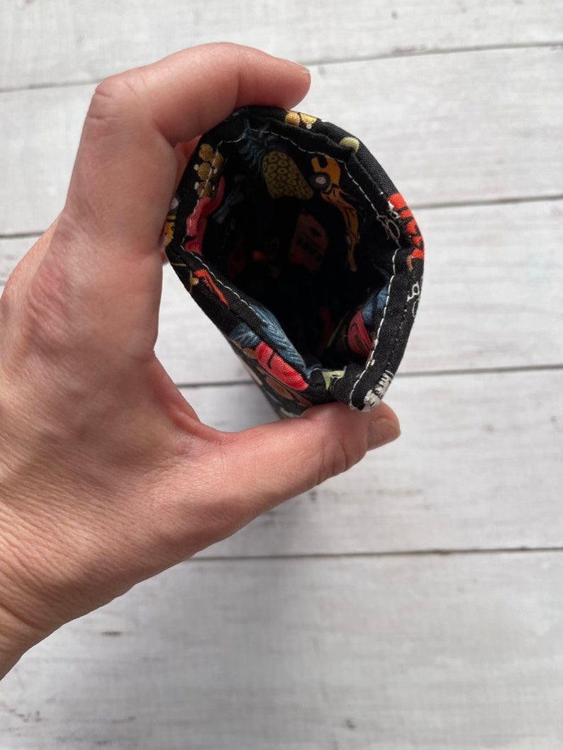 May include: A small, open fabric pouch held in a hand. The pouch is black with a colorful pattern of fruits and flowers. The interior is black. The pouch is made of a soft, flexible material.