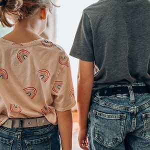 May include: Two children, a girl wearing a pink shirt with rainbow designs and a boy wearing a gray t-shirt, stand facing away from the camera. Both children are wearing blue jeans with adjustable belts.