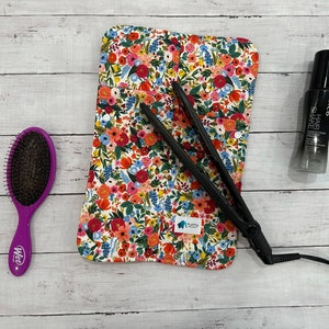 May include: A black flat iron, a purple hairbrush, and a bottle of hair product are on a white wooden surface. A floral print heat-resistant mat with a blue label is in the centre. The items are for hair styling.