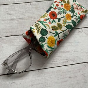 May include: A floral patterned glasses case with a pair of clear glasses resting on top. The case is made of fabric and has a white background with red, yellow, and green flowers.