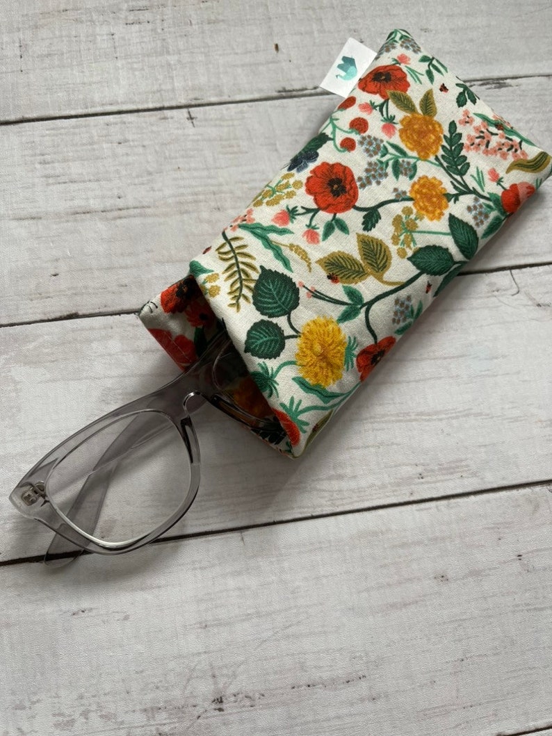 May include: A floral patterned glasses case with a pair of clear glasses resting on top. The case is made of fabric and has a white background with red, yellow, and green flowers.