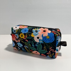 Peut inclure: Une trousse de maquillage rectangulaire à imprimé floral avec une fermeture éclair noire et une petite poignée noire. Le sac présente un fond bleu marine foncé avec un motif de fleurs roses, bleues, jaunes et blanches et de feuilles vertes.
