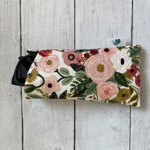 May include: A floral sunglasses case with a white background. The case is covered in a pattern of pink, red, and white flowers with green leaves. Black sunglasses are partially visible inside the case.