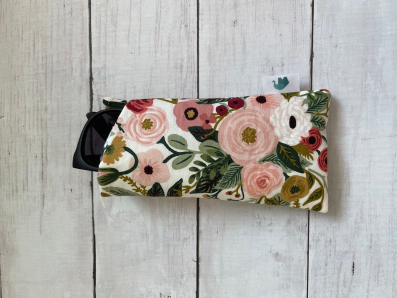 May include: A floral sunglasses case with a white background. The case is covered in a pattern of pink, red, and white flowers with green leaves. Black sunglasses are partially visible inside the case.