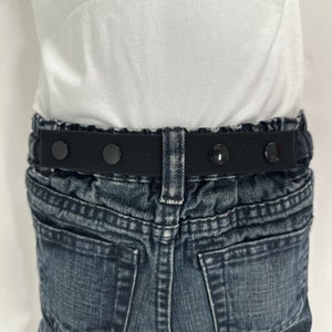 May include: Black elastic belt with four adjustable buttons, worn over a pair of blue jeans.