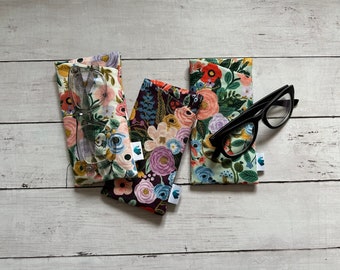 May include: Three floral patterned eyeglass cases with a pair of clear glasses and a pair of black glasses on a white wooden surface.