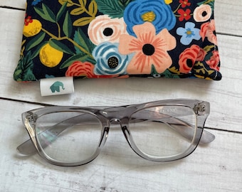 May include: A pair of grey glasses with clear lenses and a floral print eyeglass case. The case has a dark blue background with pink, red, blue and yellow flowers.