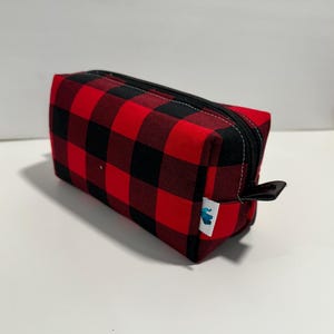 Peut inclure: Une trousse rectangulaire à motif de carreaux rouges et noirs. La trousse est dotée d'une fermeture éclair noire et d'une petite étiquette en tissu avec un motif bleu. La trousse est en tissu doux.