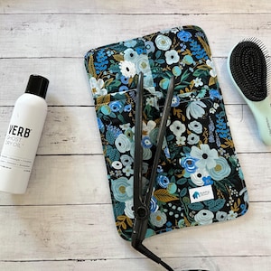 May include: A flat iron, hairbrush, and hair product are arranged on a light wood surface. The flat iron is black. The hairbrush is light green with black bristles. The hair product is a white bottle with black text that reads "VERB GHOST DRY OIL". A floral print case holds the flat iron.