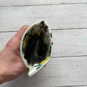 May include: A white fabric pouch with a floral print in shades of green and yellow. The pouch is open and contains a pair of black sunglasses.