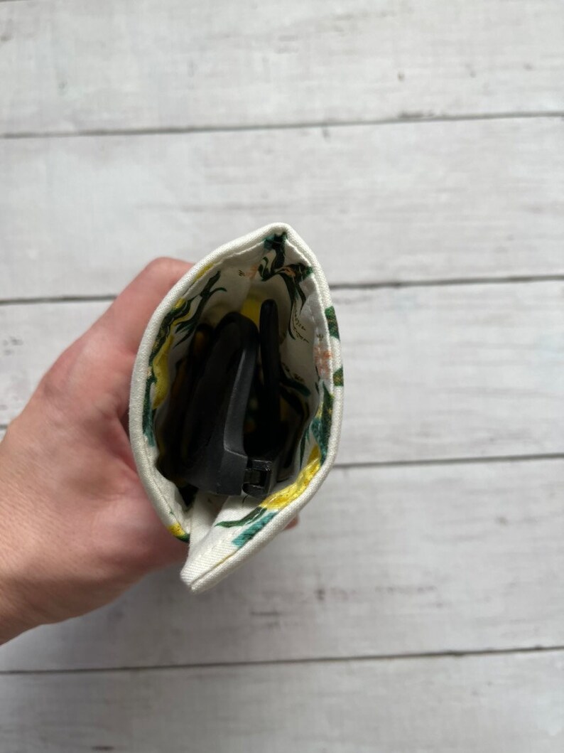 May include: A white fabric pouch with a floral print in shades of green and yellow. The pouch is open and contains a pair of black sunglasses.
