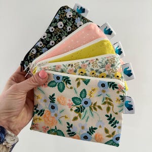 May include: A hand holds a stack of six small zippered pouches. The pouches display floral patterns in black, pink, yellow, and cream. Each pouch has a white tag with a blue and teal design.
