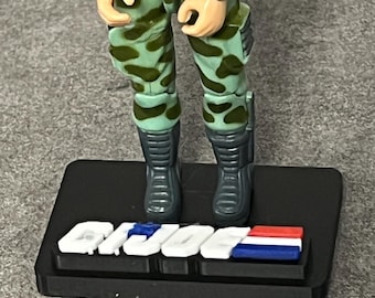 May include: A plastic action figure wearing light green camouflage pants and dark gray boots, standing on a black rectangular display base. The base features the white, blue, and red G.I. JOE logo text embossed on the front surface.