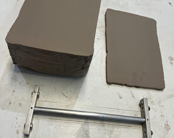 May include: A large rectangular block of gray clay, a thinner rectangular slab of gray clay, and a metal wire clay cutter tool with a horizontal bar and two vertical handles, used for slicing and shaping pottery materials in a studio.