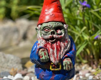 May include: A zombie garden gnome figurine featuring a pointed red hat, a blue jacket, and brown boots. The gnome has pale green skin, a white beard stained with red blood-like paint, and one protruding eye, with red splatters across its clothing.