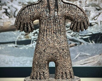May include: A brown and black speckled stone-like figurine of a humanoid creature with outstretched arms, wide feet, and two vertical lines extending from the eyes down the face, all mounted on a circular base with a textured, granular surface.