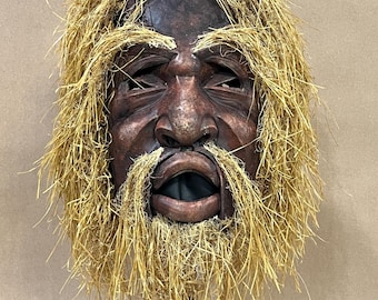 May include: A dark brown carved wooden mask featuring a human face with an open mouth, deep-set eyes, and a prominent nose. The mask is adorned with a thick, shaggy mane and beard made of light yellow straw-like fibers covering the head and chin.