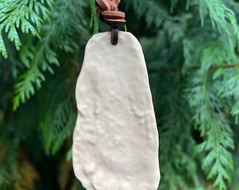 May include: A cream-colored, irregular, organic-shaped pendant with a subtle topographical map texture on its surface. The pendant is suspended from a brown leather cord that is knotted at the top, creating a simple and functional necklace design.