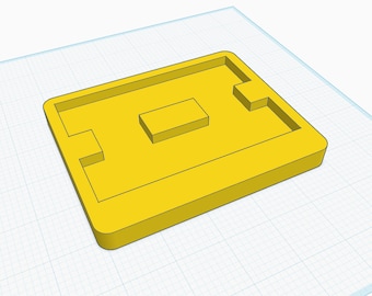 May include: A yellow plastic mold with a rectangular shape and a smaller rectangular cutout in the center. The mold has a raised edge around the perimeter.