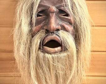 May include: A carved wooden mask featuring a dark brown finish with deep-set eyes and an open mouth. The mask is adorned with long, light-colored, fibrous hair that covers the top of the head and forms a thick, flowing beard around the chin.