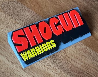May include: A rectangular sign featuring the text SHOGUN in bold red letters and WARRIORS in yellow letters below it. The text is mounted on a black background, which is set against a light gray base with a textured surface finish.