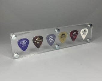 May include: A clear acrylic display stand holding six guitar picks in a row. The picks include a purple Dunlop 2.0 mm, a brown Jim Dunlop X-Heavy, a purple Dunlop Gator, a gold Dunlop Ultex 1.0, a red Jim Dunlop Heavy, and a white Dunlop Sharp 1.5 mm.