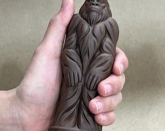 May include: A dark brown, solid-colored figurine depicting a standing, ape-like creature with a textured, fur-like surface. The figure features a defined face with a prominent brow, a beard, and long arms resting at its sides, all set upon a flat base.