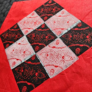 May include: A square red fabric patch featuring a central diamond-shaped grid of alternating black and light pink squares. Each square is decorated with a detailed black ink print of spiders and web patterns, creating a repeating graphic design.