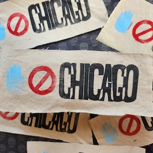 May include: Several tan fabric patches with the word "CHICAGO" printed in black. Each patch also features a red prohibition symbol, and a blue handprint. The patches are scattered on a dark background.