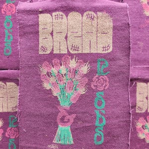 May include: A rectangular purple fabric patch featuring the word BREAD in large tan letters at the top. Below the text is a graphic of a bouquet of pink roses with green stems and leaves. The word ROSES is printed vertically in green on the right.