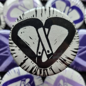 May include: A circular pinback button featuring a high-contrast black and white graphic of two interlocking carabiners forming a heart shape. The design includes textured, ink-like radiating lines extending from the centre towards the outer edge.