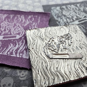 Op de afbeelding: Een vierkante linoleum stempel met een uitgesneden ontwerp van een sigaret met wervelende rook, vergezeld door een paarse stoffen patch en een zwarte stoffen patch, beide met hetzelfde gedrukte ontwerp van een sigaret en rook.