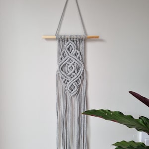 May include: A grey macrame wall hanging with a geometric design and long fringe. The macrame is suspended from a natural wood dowel and a grey cord, creating a triangular shape. The wall hanging is displayed against a light grey wall.