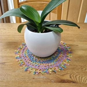 May include: A white ceramic pot with a green plant rests on a colourful, hand-crocheted doily. The doily has a circular design with pastel colours, including blue, yellow, and purple. The plant has long, green leaves. The items are on a wooden table.