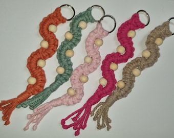May include: Five macrame keychains in shades of orange, green, pink, fuchsia, and tan. Each keychain features a wavy design with wooden beads and a metal ring for attaching keys. The ends are finished with a tassel.