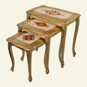 Può includere: Un set di tre tavolini a incastro con telai color oro e gambe curve. Ogni tavolino presenta un piano rettangolare con un pannello centrale color crema decorato con un motivo floreale rosso e oro, circondato da un bordo dorato testurizzato.